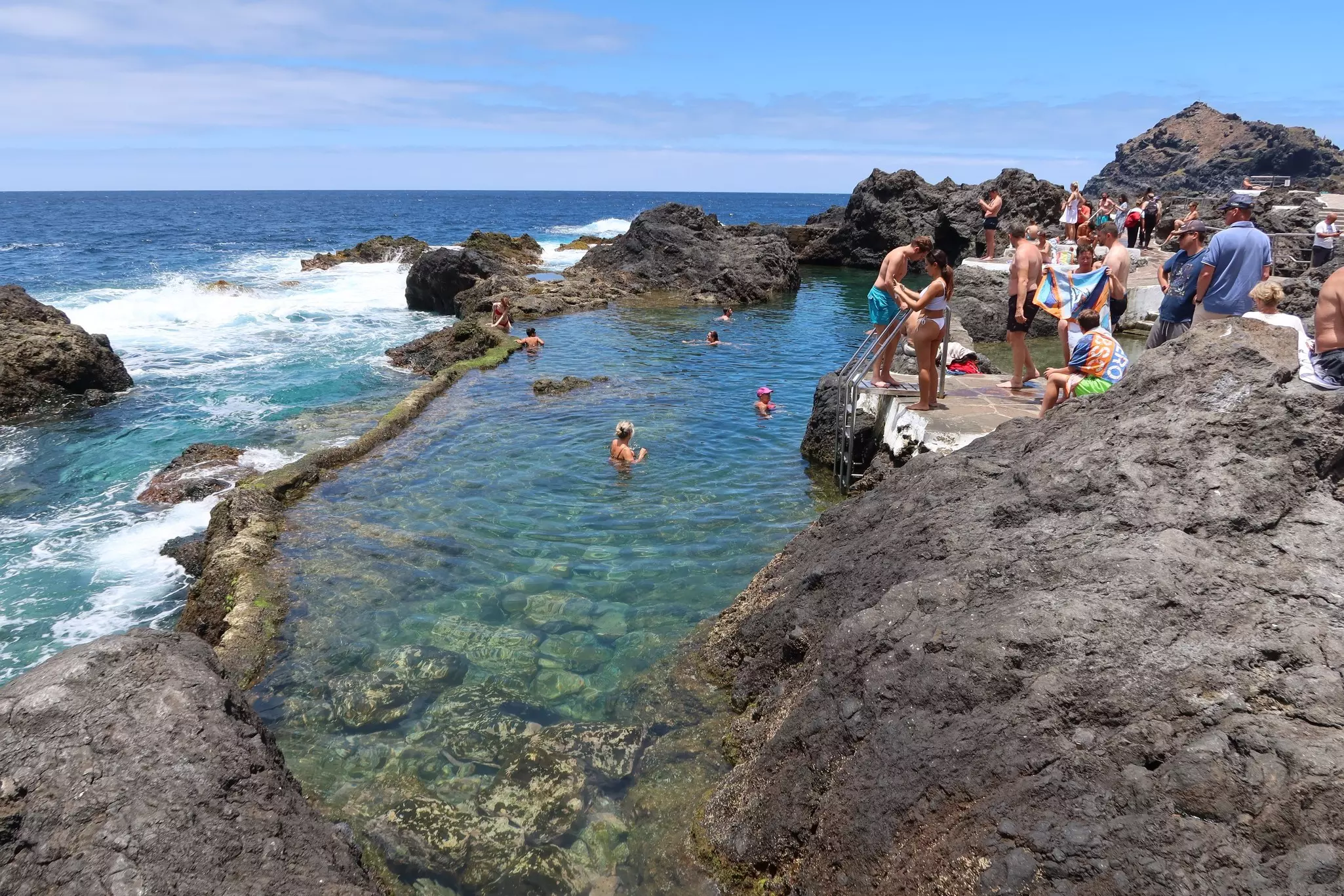 People swim in tidal pools next to the crashing surf on a shoreline with volcanic rocks
