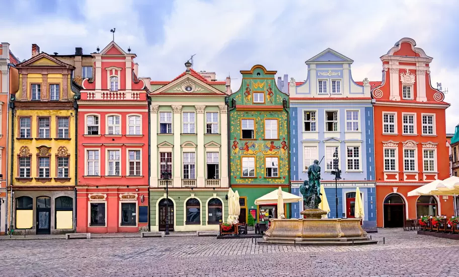 Row of tall, colourfully painted townhouses facing a cobbled square in Poznań, with a bronze Neptune fountain in front.