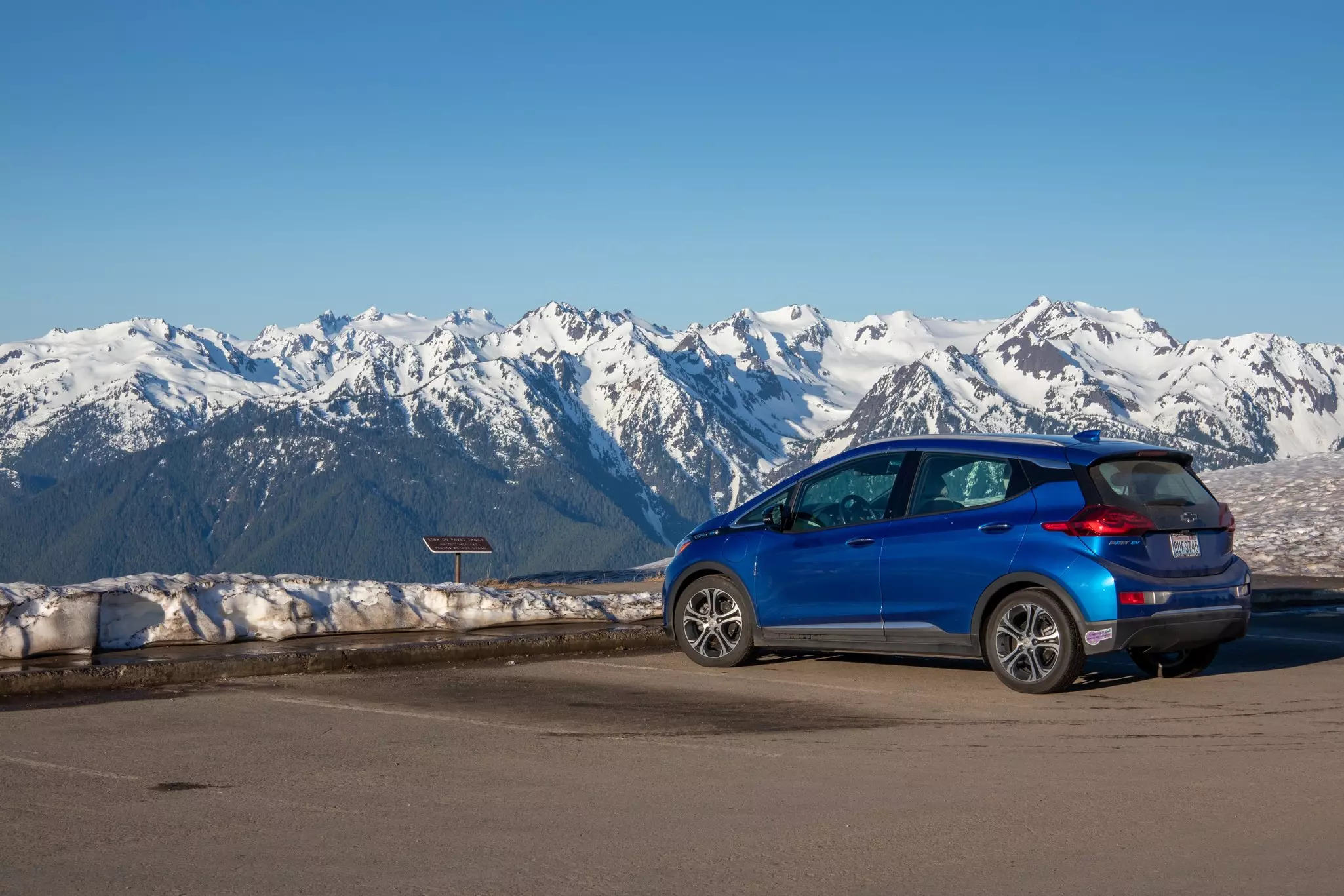 A blue car parked at an overlook facing snow-covered mountains.