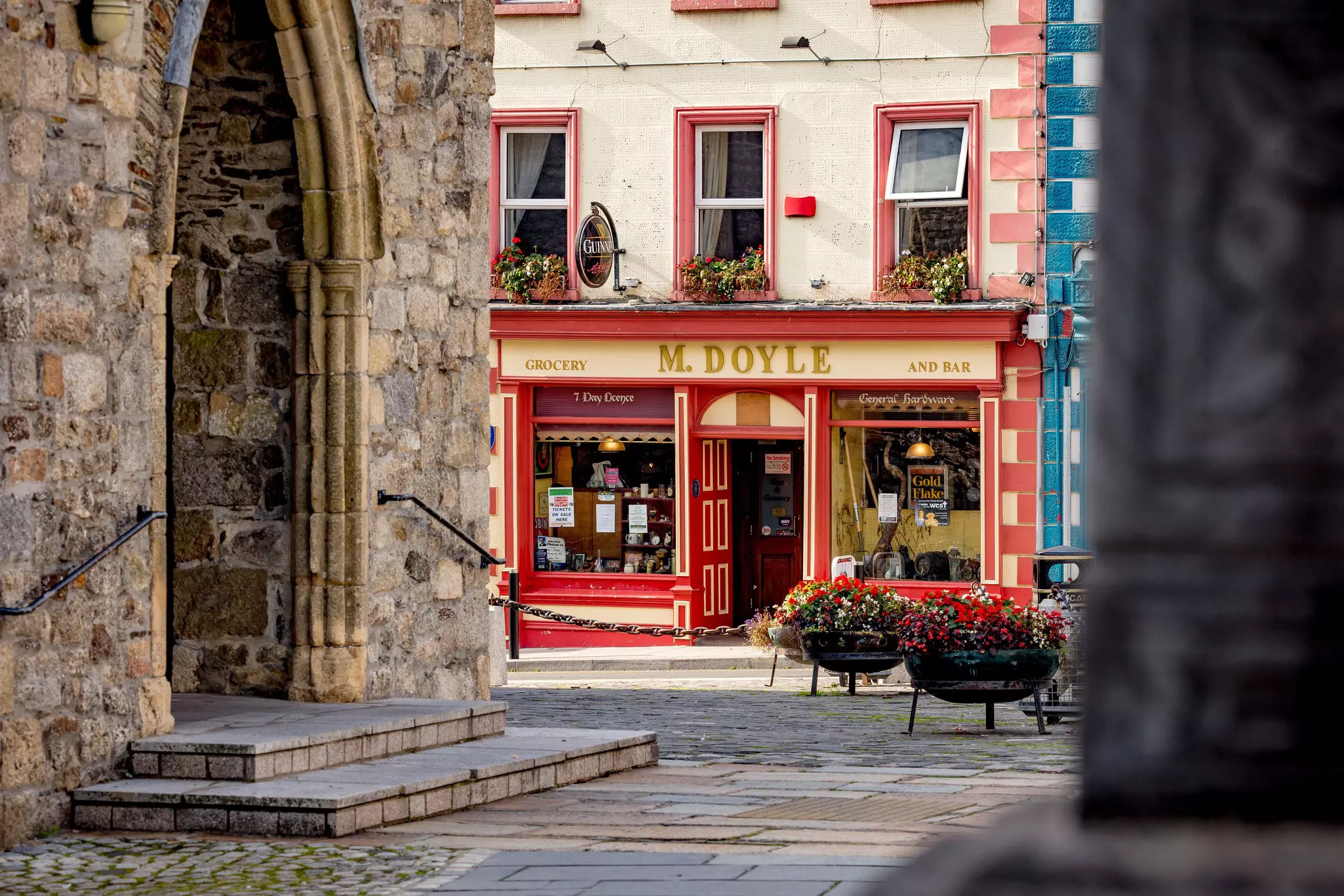 The exterior of a traditional Irish pub. The pub is framed by rows of flower boxes and to the left is the entrance of a stone church.