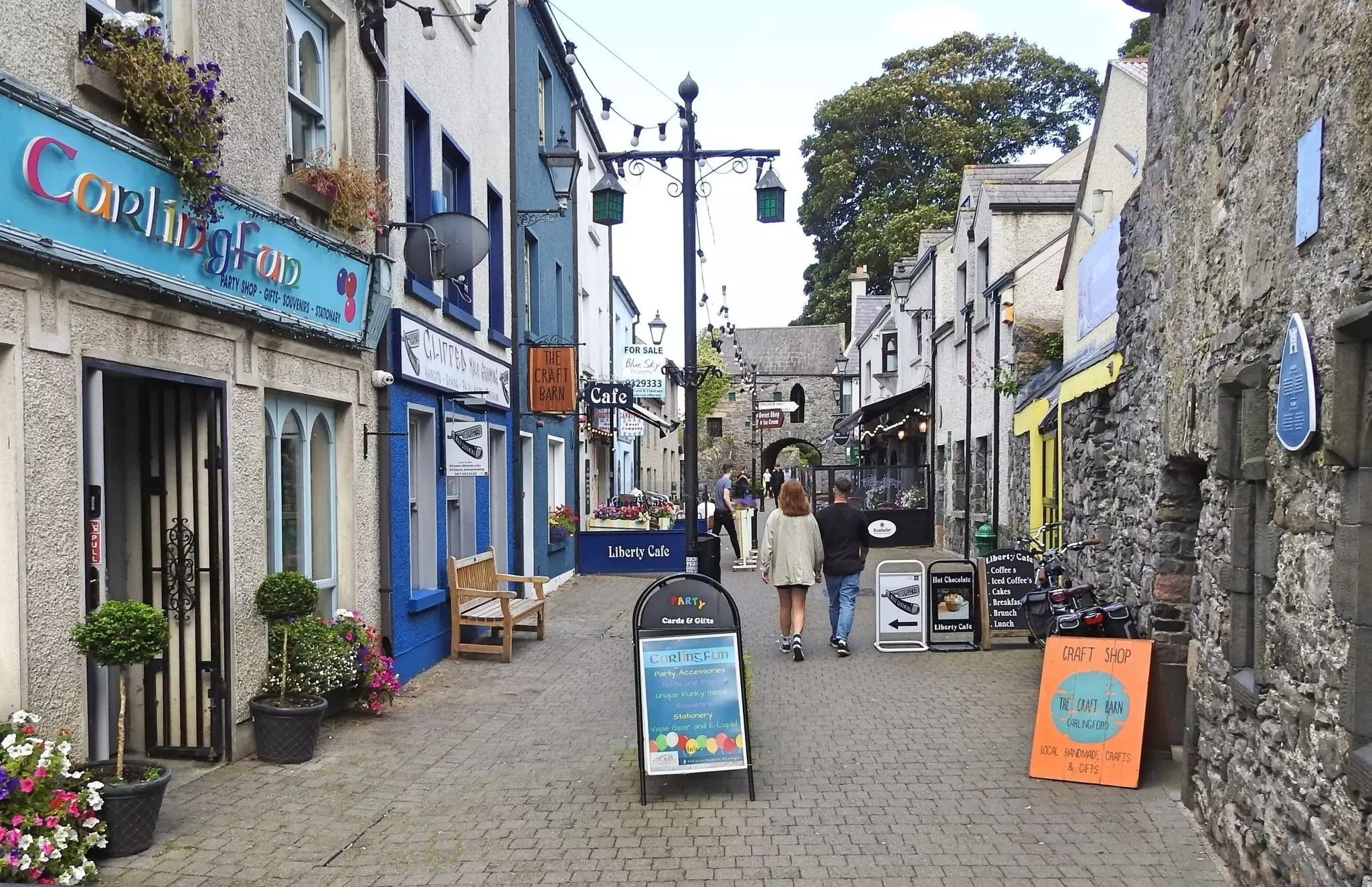 People walk along a pedestrianized cobblestone street lined with craft shops and cafes.