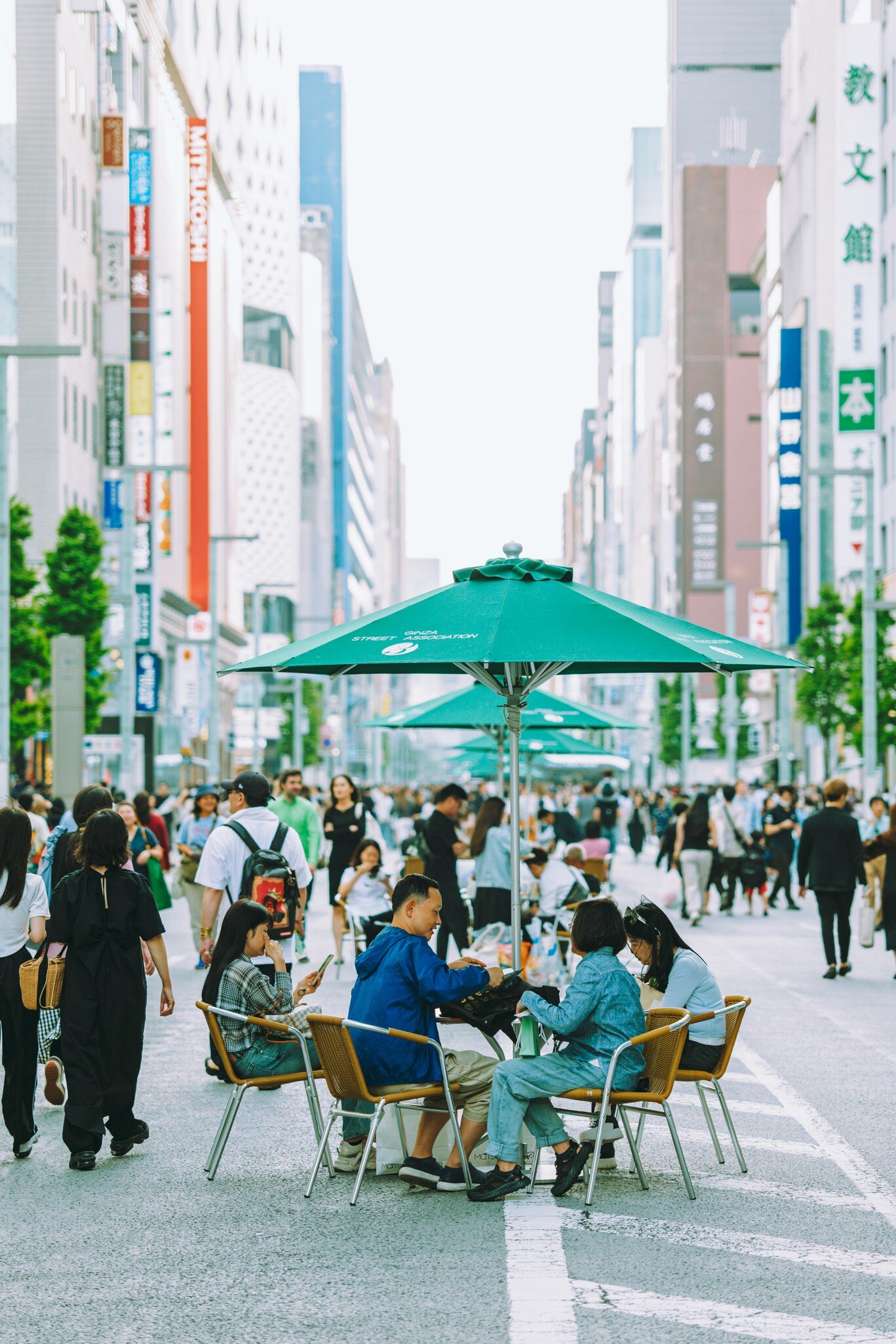 Crowds walking and sitting at tables under green umbrellas in the middle of a wide Ginza avenue lined with tall store signs.