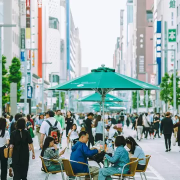 Crowds walking and sitting at tables under green umbrellas in the middle of a wide Ginza avenue lined with tall store signs.