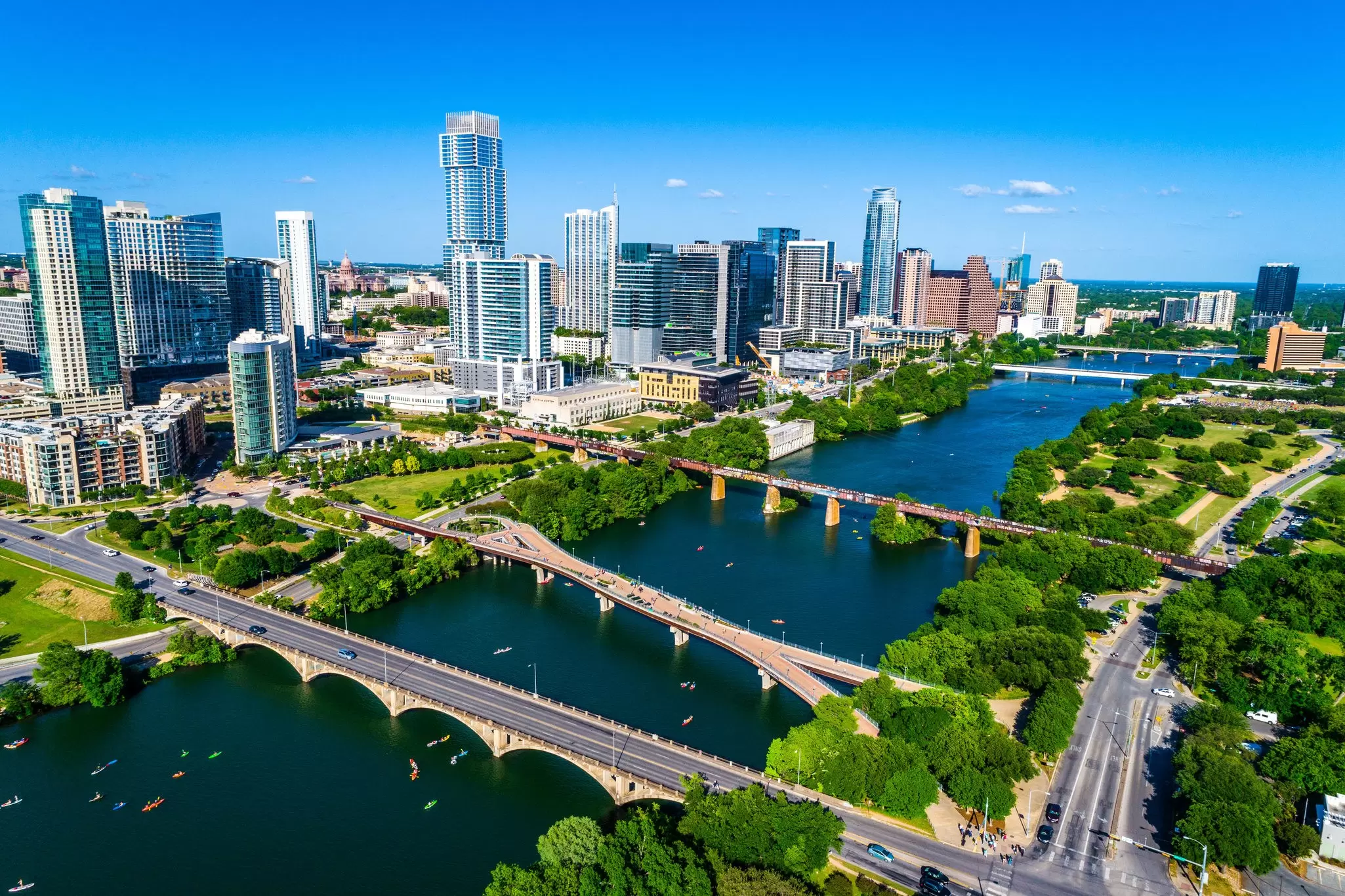 Aerial view of the Austin skyline beside a river lined with green parkland and crossed by bridges, with kayaks on the water.