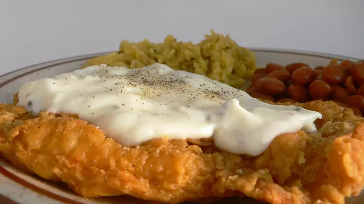 A close up image of fried meat with gravy, rice and beans.