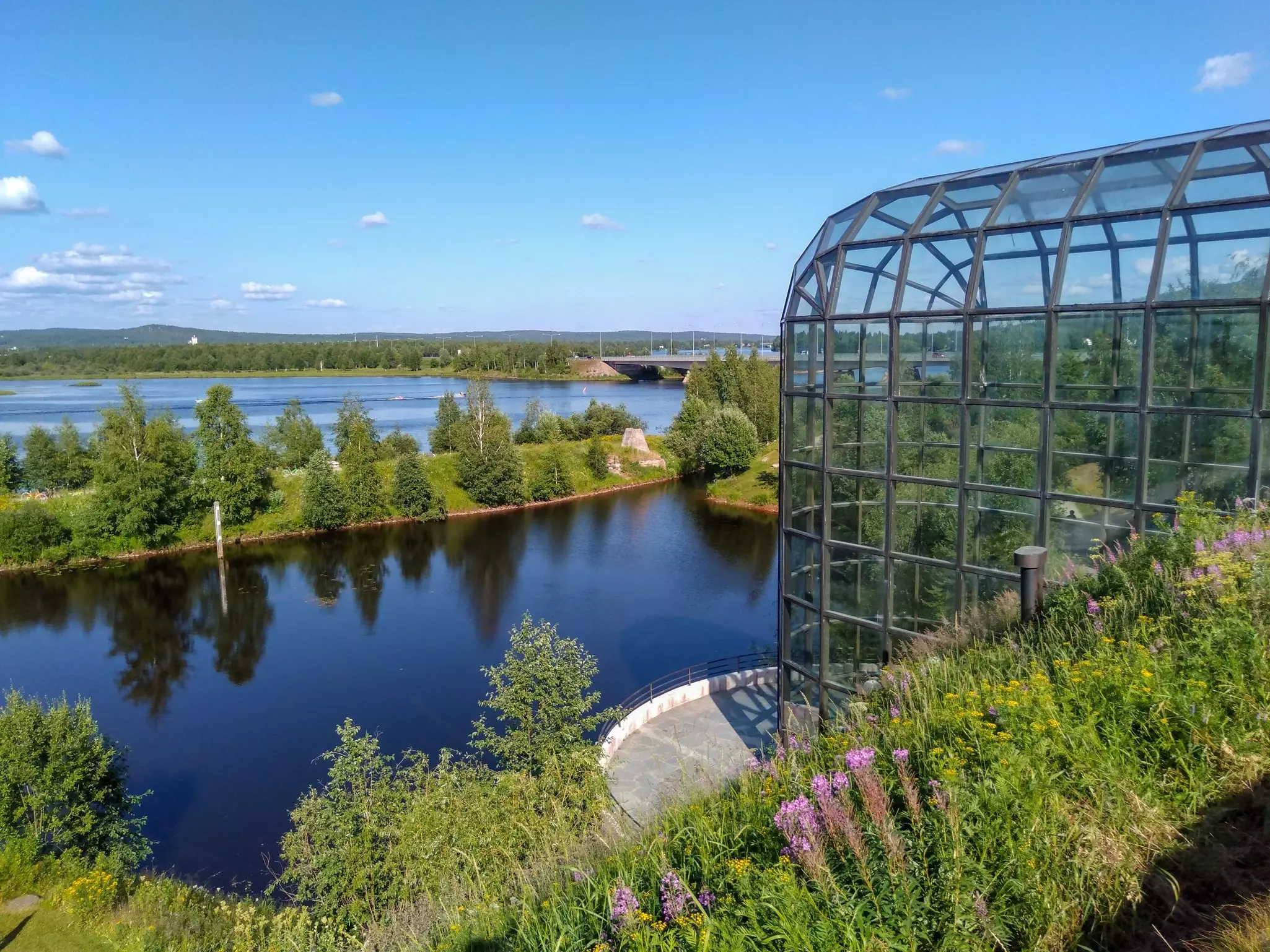 A glass and metal framed atrium is next to a small inlet of a larger lake surrounded by trees.