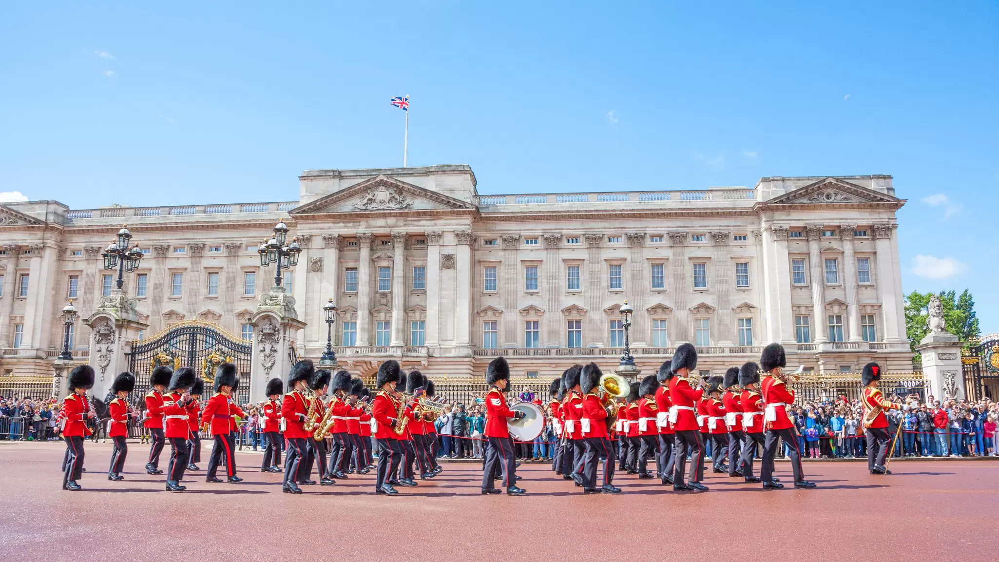 A row of soldiers in red uniforms with tall black hats march in front of a white/grey palace building