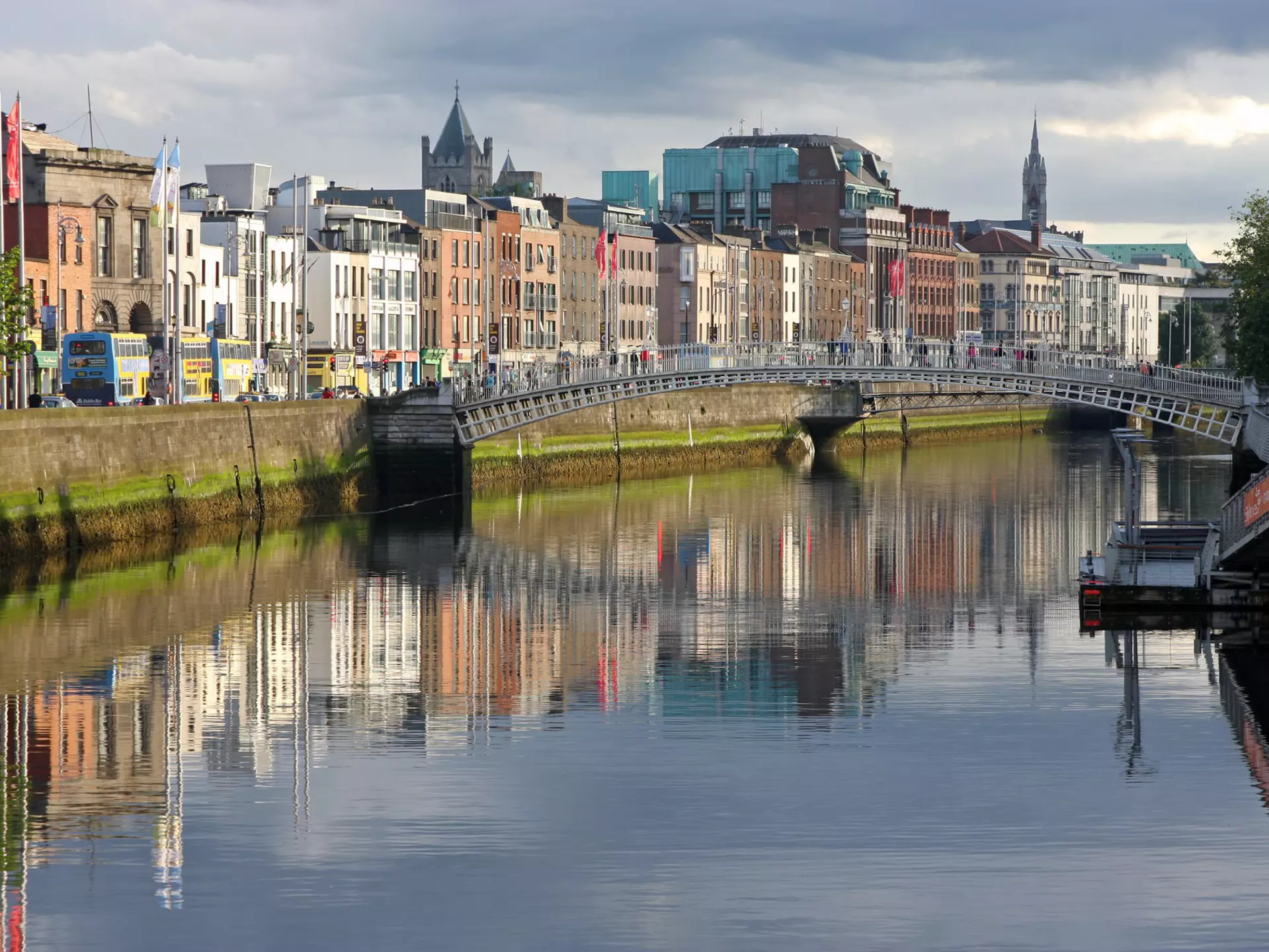 A pedestrian bridge crossing a river. Buildings lining the waterfront are reflected in the water.