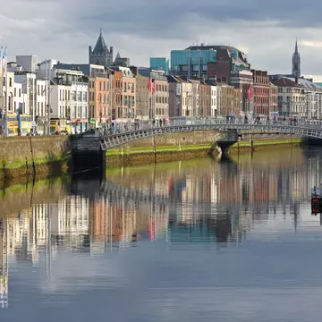 A pedestrian bridge crossing a river. Buildings lining the waterfront are reflected in the water.