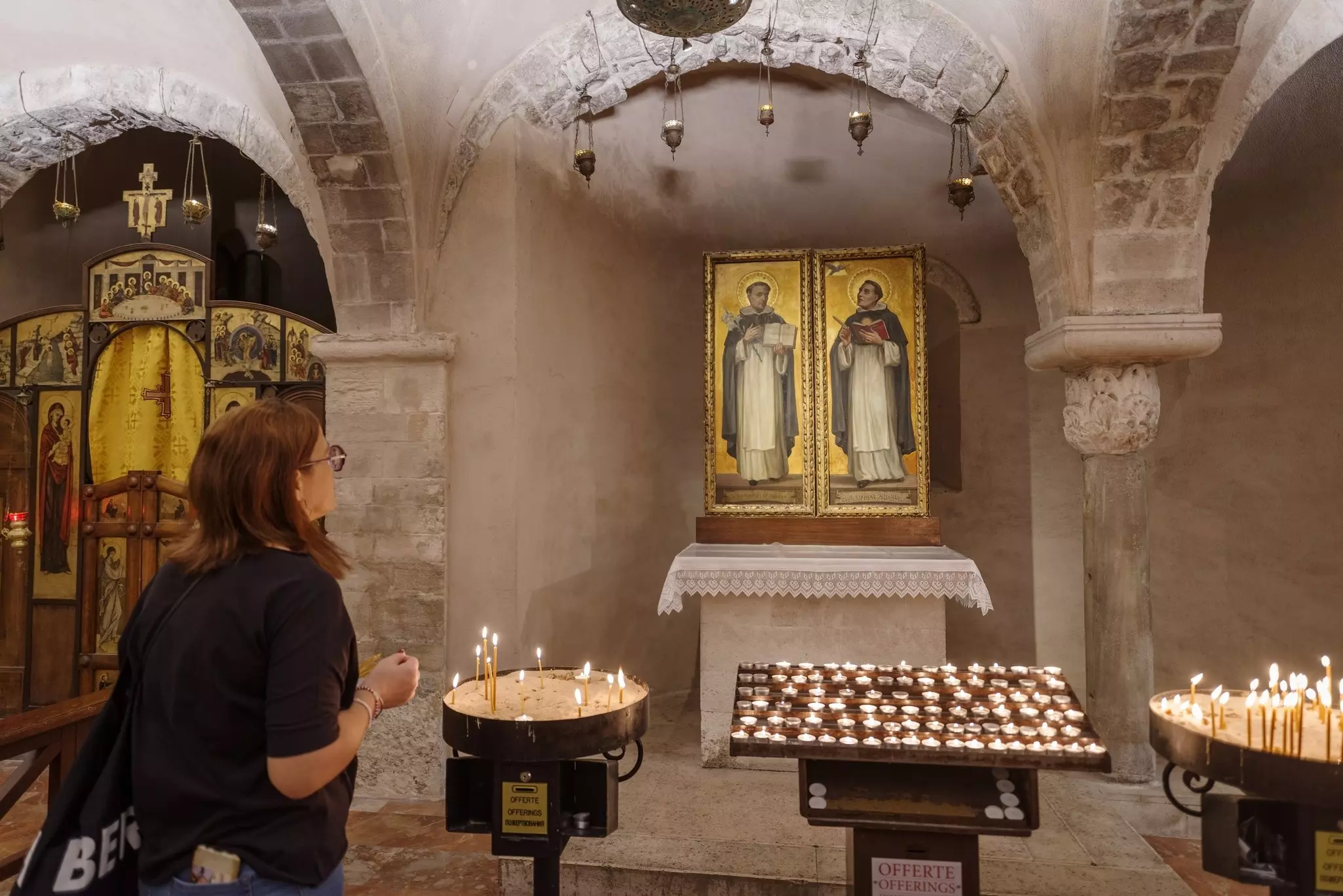 A person stands in front of votive candles by a painting of saints in the crypt of a church.