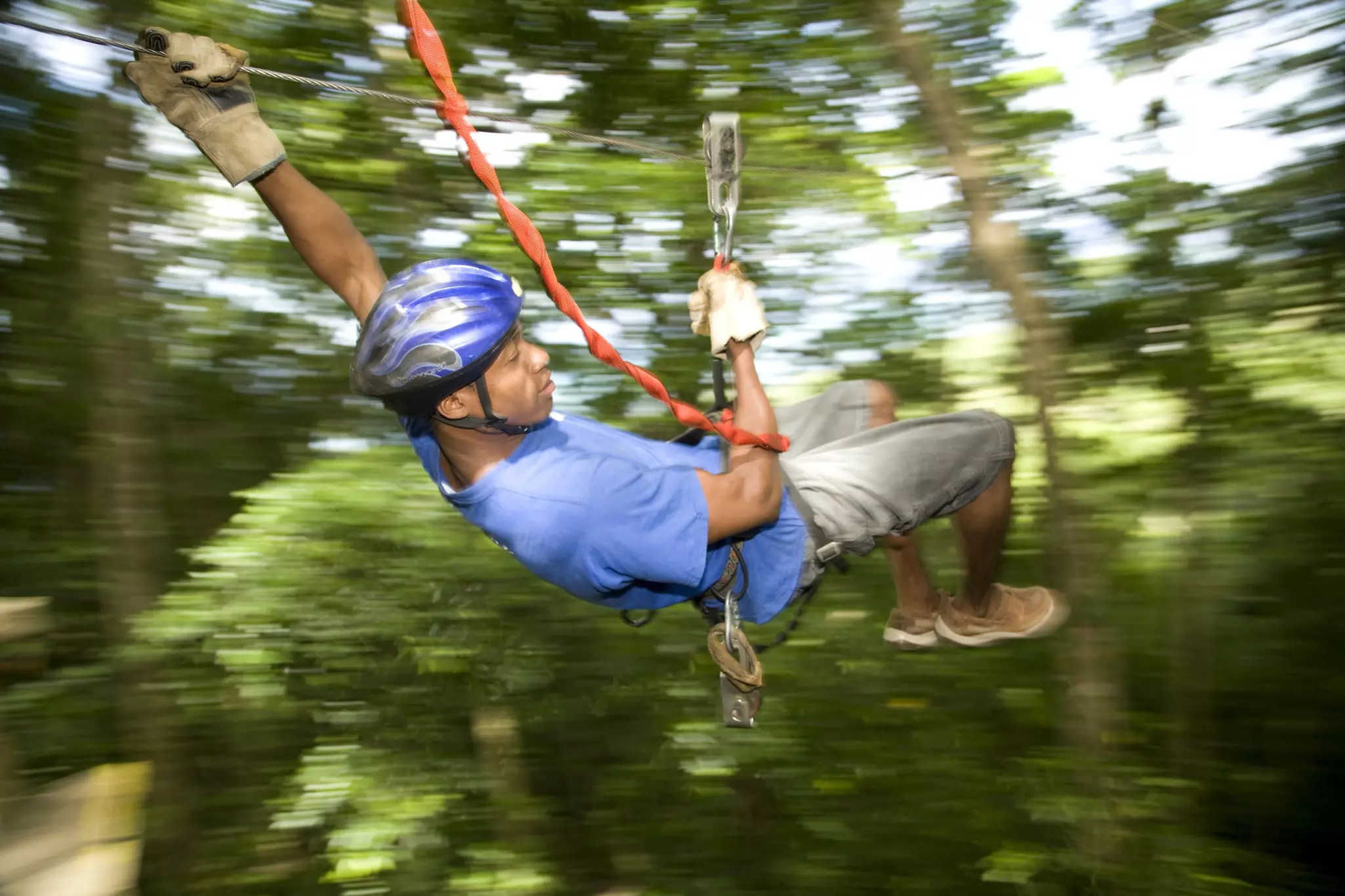 A man grips a harness as he whisks down a zip line in a jungle. The blurry background indicates his speed.