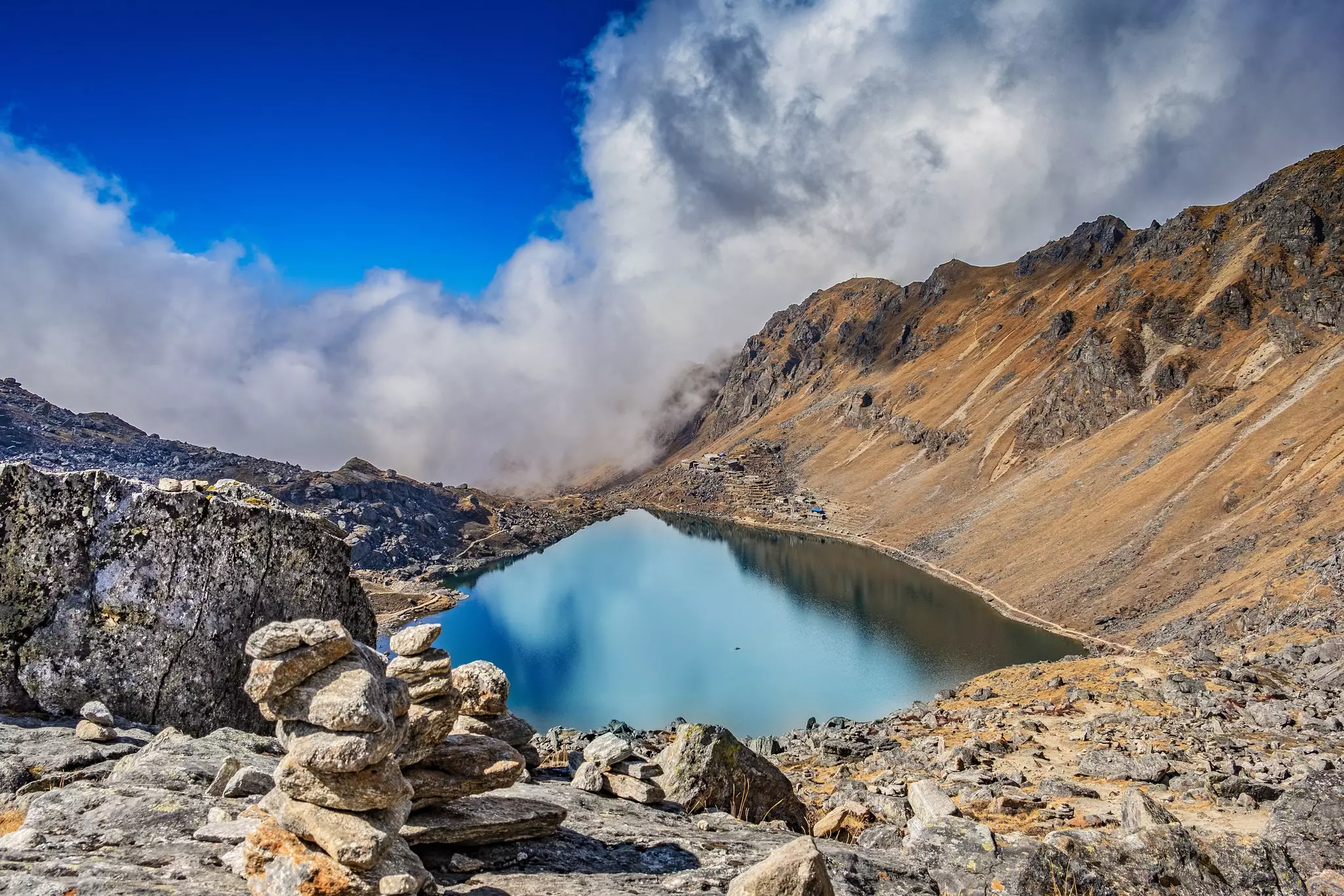 A view from above of a glassy, high-altitude lake. Rocky slopes lead to its shores, with clouds hanging low in the sky.