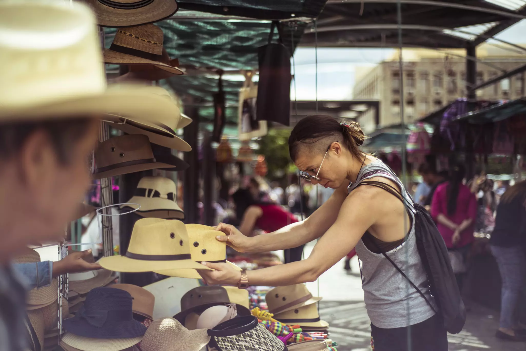 A person wearing in sunglasses examines a display of brimmed hats at an open-air market