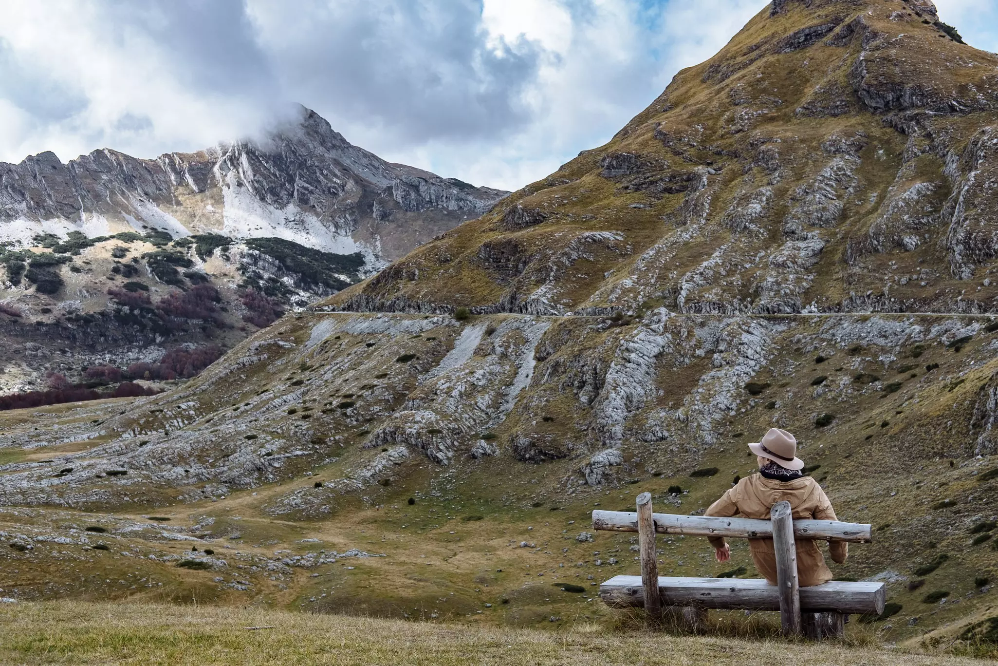 A person sits on a rustic log bench in front of grassy and rocky mountains covered in a light frost.