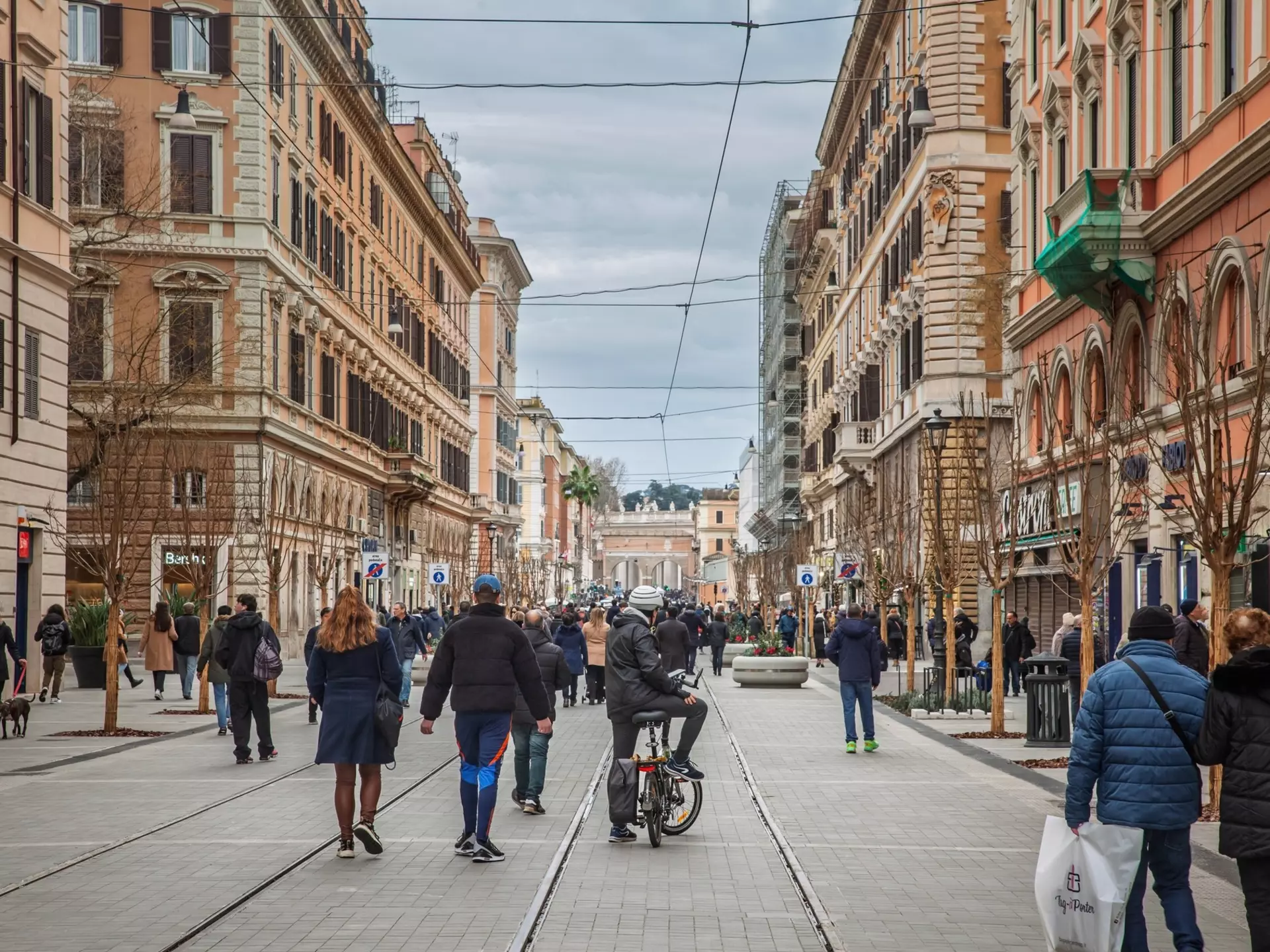 People walking on a busy street with cyclists and tram lines. Historic facades and shops frame pedestrians.