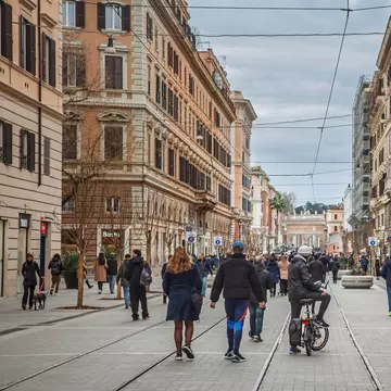 People walking on a busy street with cyclists and tram lines. Historic facades and shops frame pedestrians.