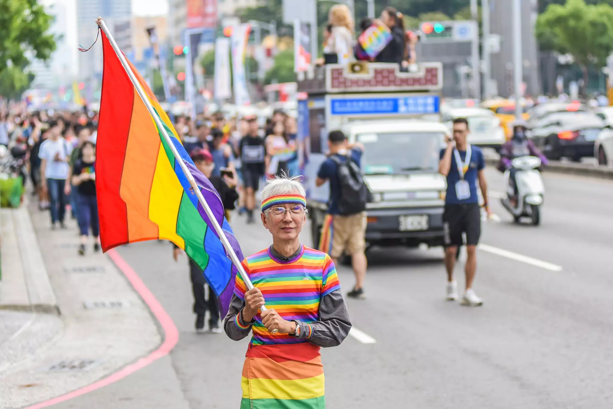 A man wearing a rainbow shirt carries a rainbow flag in a parade.