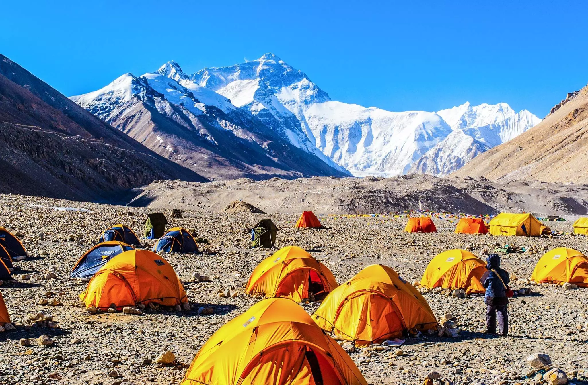 A series of yellow tents pitched on a rocky terrain at the foot of a snow-capped mountain
