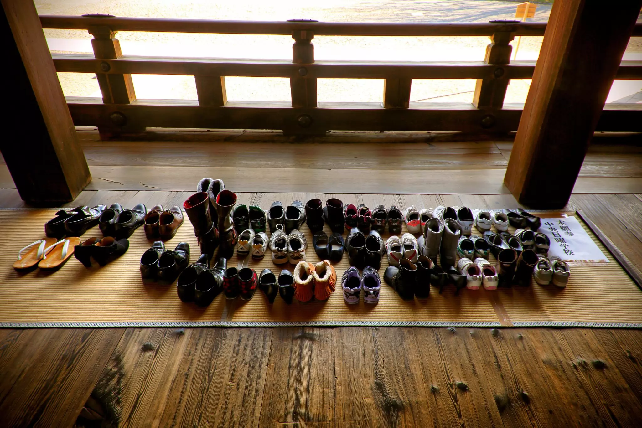 A view from above of dozens of pairs of shoes on a mat laid on a wooden floor. Two columns and a railing are seen next to the mat.