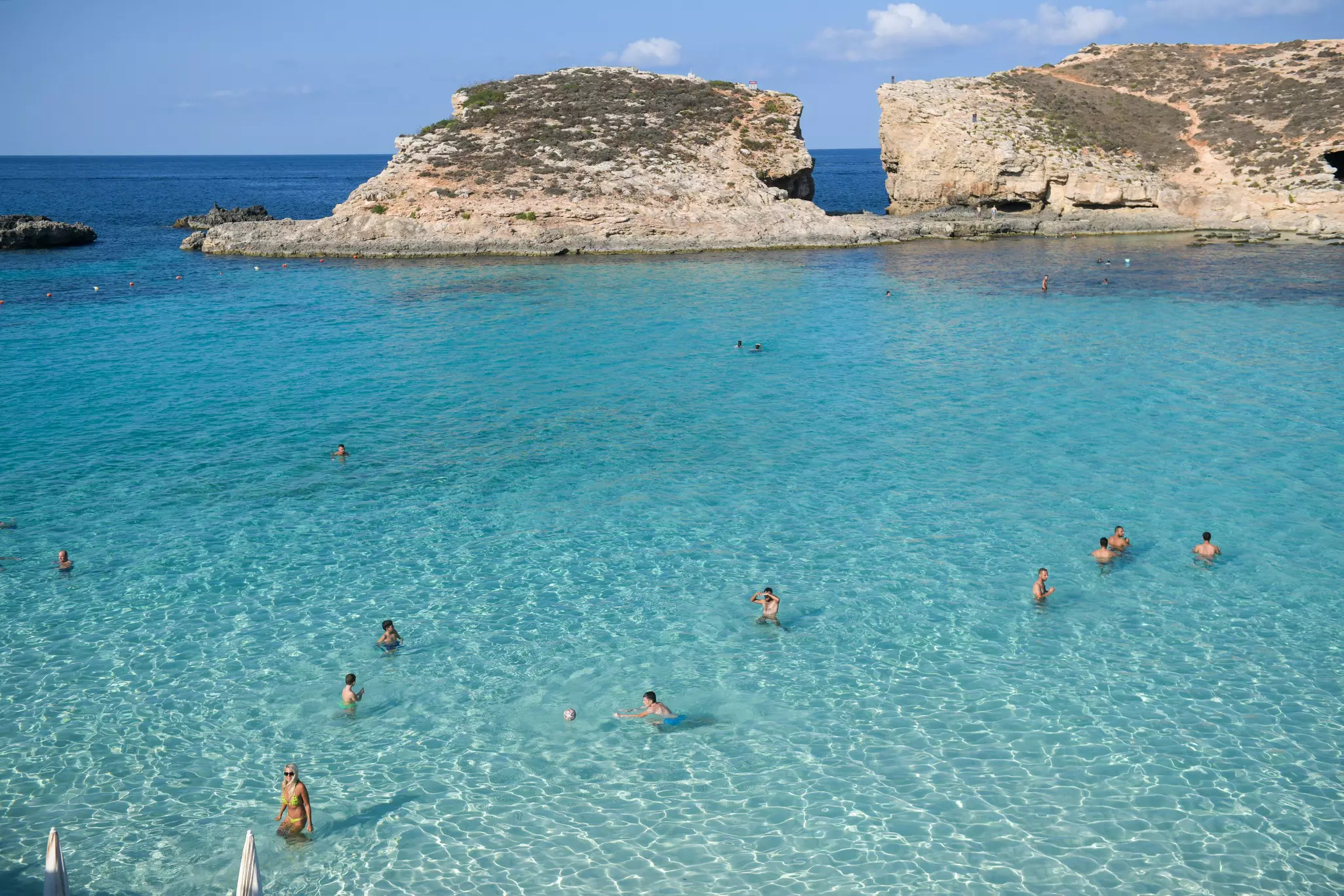 People swim in a bright blue lagoon with large rocks in the distance on a sunny day.