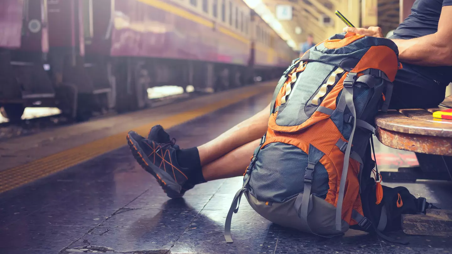A man is sitting on a bench at a train station and looking at his phone, There is a large backpack sitting on the floor beside him, and a purple train is stationary at the platform.