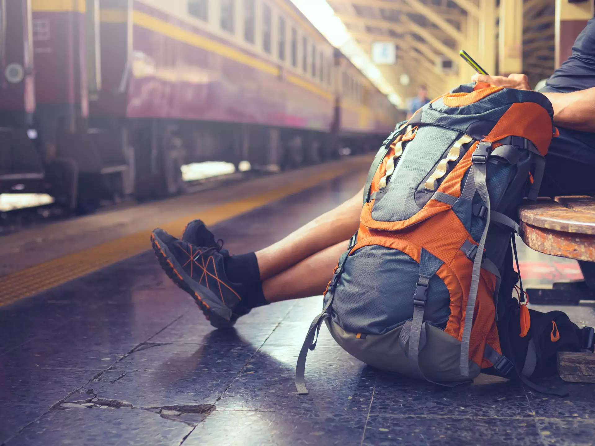 A man is sitting on a bench at a train station and looking at his phone, There is a large backpack sitting on the floor beside him, and a purple train is stationary at the platform.