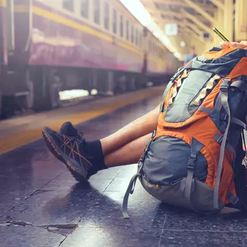 A man is sitting on a bench at a train station and looking at his phone, There is a large backpack sitting on the floor beside him, and a purple train is stationary at the platform.