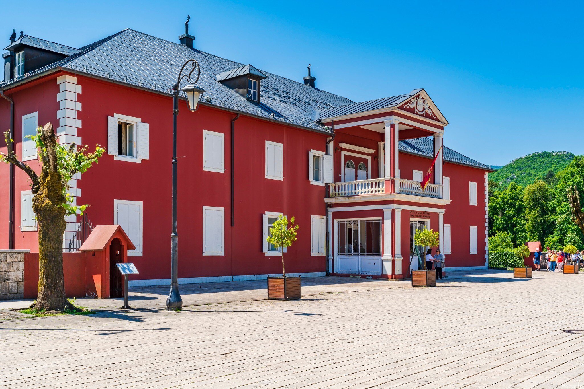 Two-storey red building with white shutters, a columned balcony portico and a flag, visitors on the paved square