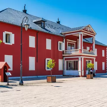 Two-storey red building with white shutters, a columned balcony portico and a flag, visitors on the paved square