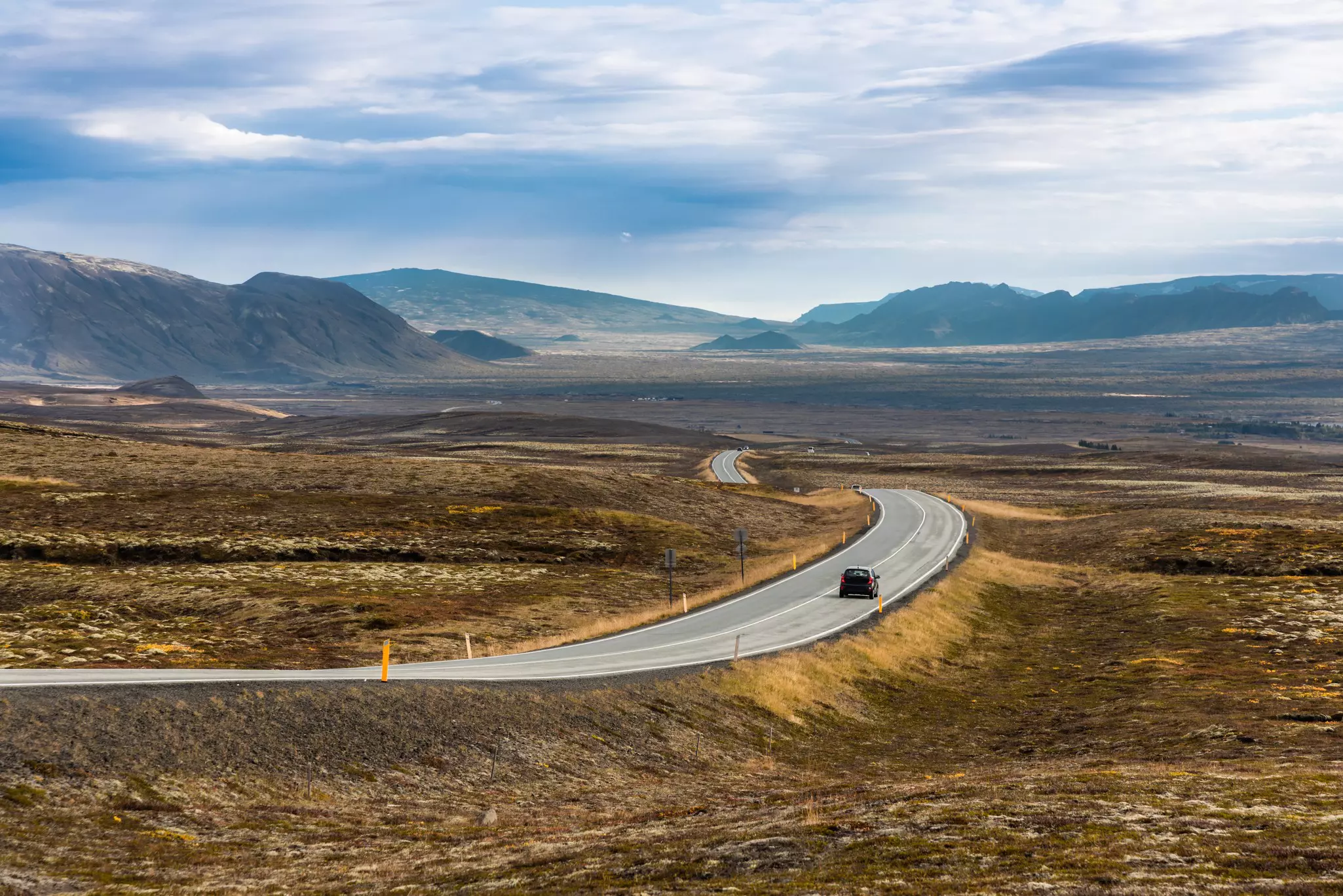 A small car drives along a main road cutting through a volcanic landscape.
