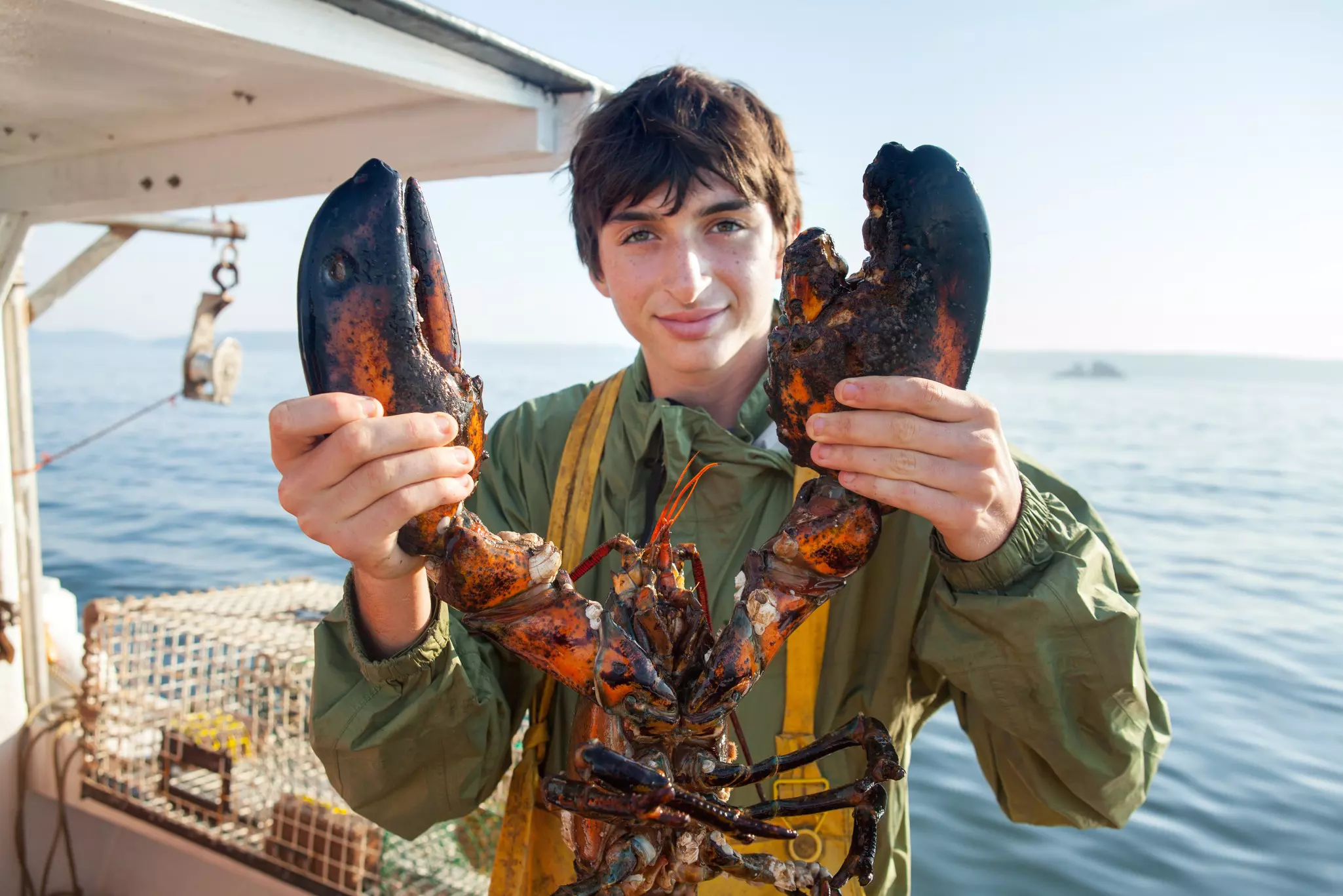 Young man wearing yellow waders and a green jacket holds up a lobster while standing on the deck of a boat, with a white lobster cage and the ocean behind him