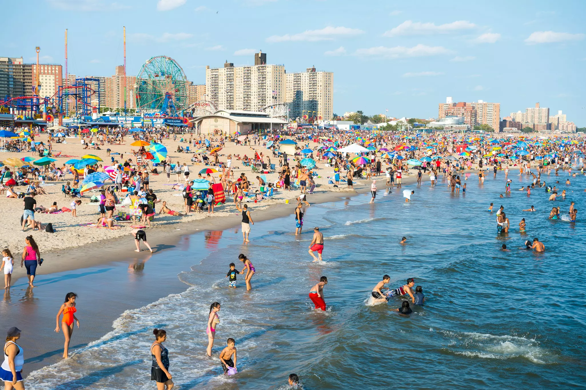 Crowds of people sunbathing and swimming at beach with an amusement park.