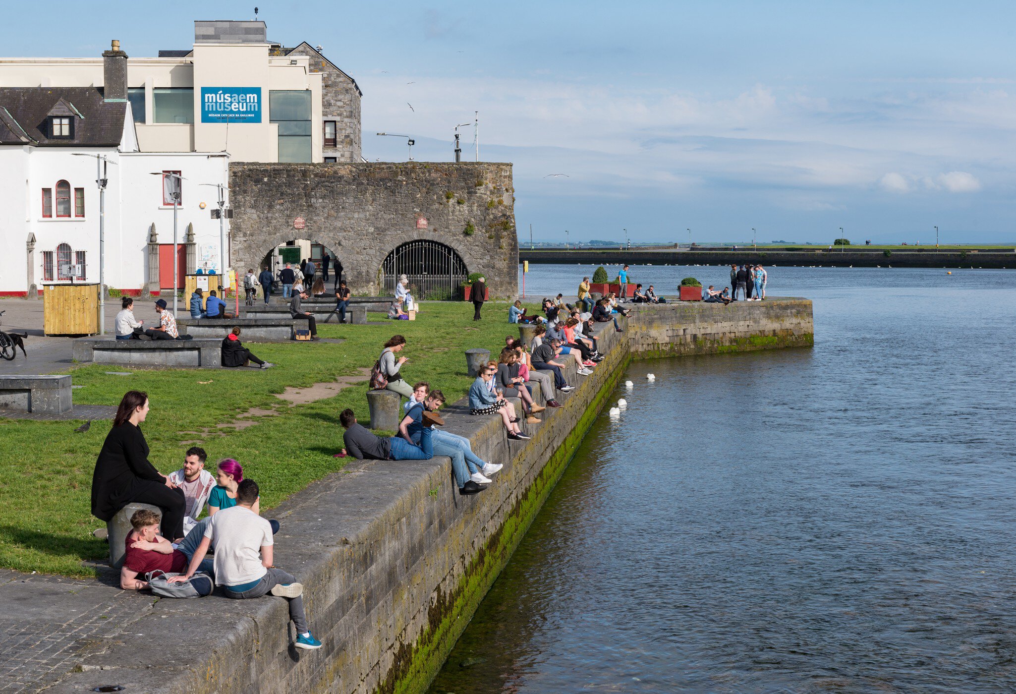  People socialising and enjoying the view from Spanish Arch along the bank of Corrib river in Galway city, Ireland.  A row of people sitting with blue sky and blue water.