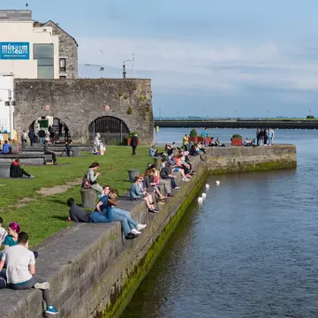  People socialising and enjoying the view from Spanish Arch along the bank of Corrib river in Galway city, Ireland.  A row of people sitting with blue sky and blue water.