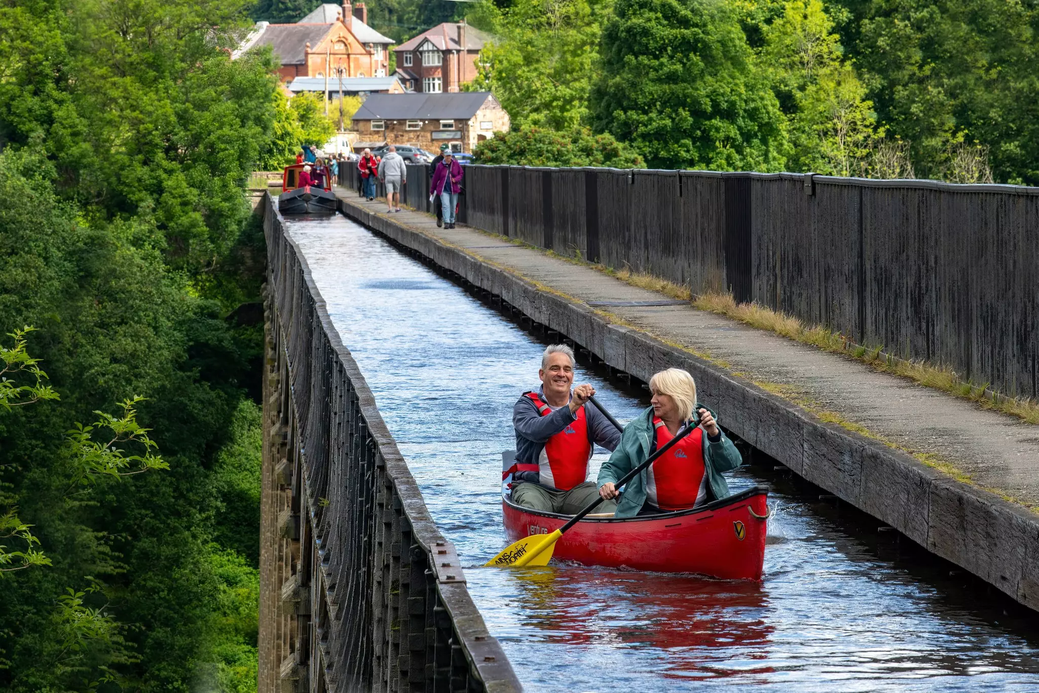 Close up of two people in a canoe crossing the Pontcysyllte Aqueduct with a canal narrowboat and people walking.