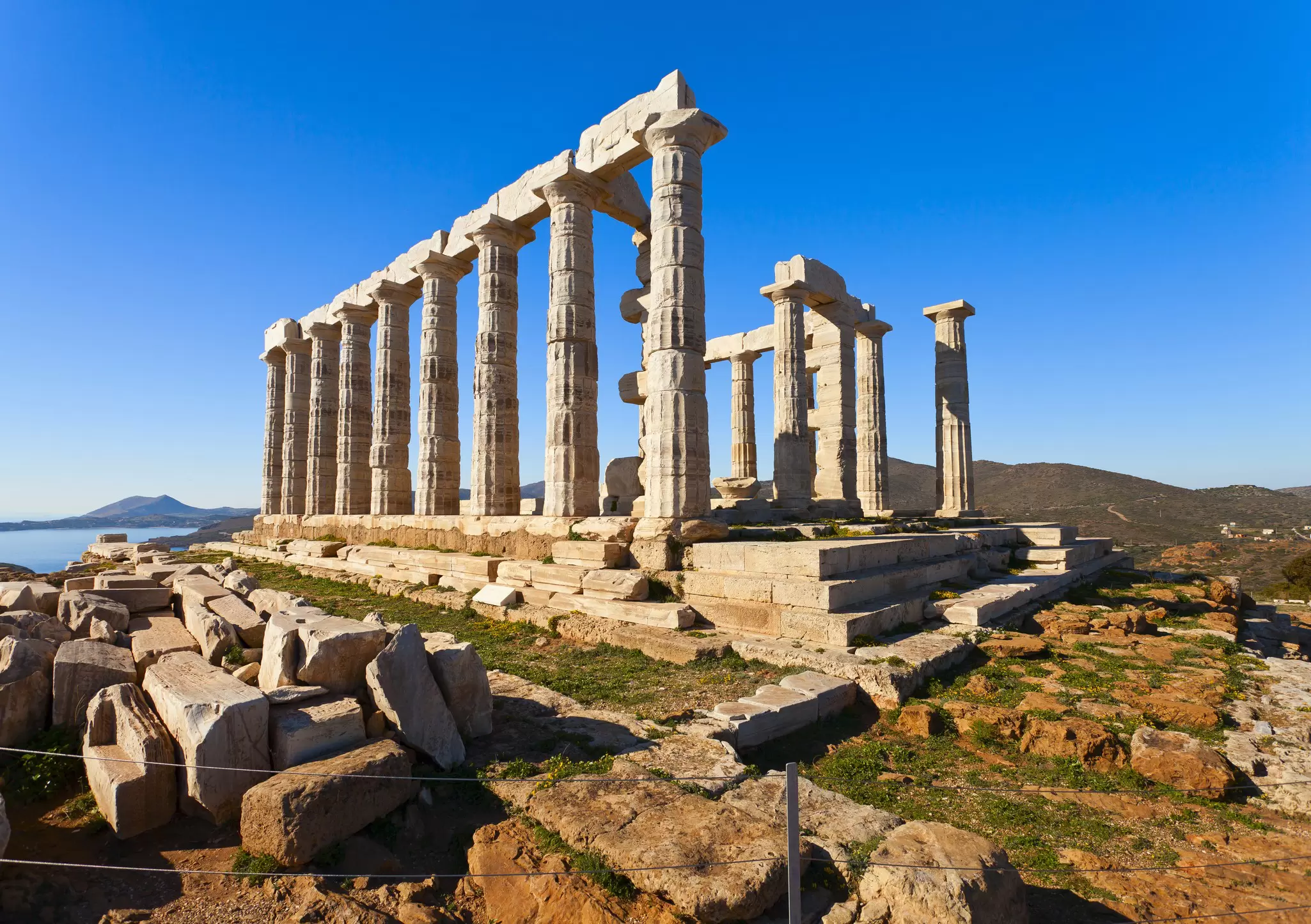 Marble Doric columns of the Temple of Poseidon stand on a rocky cape above the sea under a clear blue sky.