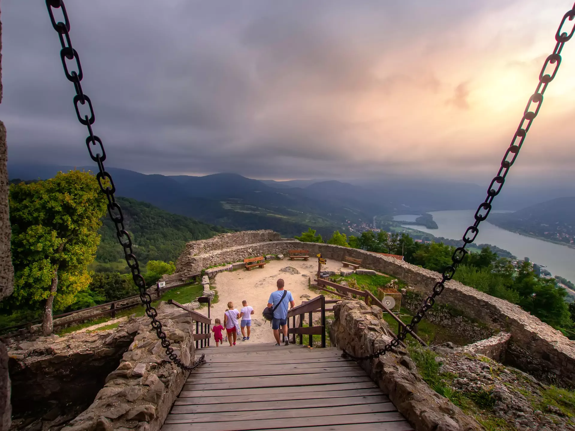 People walk down a ramp toward an overlook far above a river; a stone wall surrounds the overlook, and there are low stone walls leading to the ramp, with chains connected that rise to a point out of view.