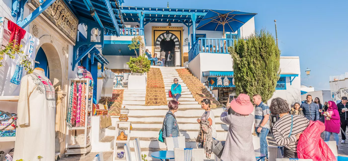A crowded square with whitewashed buildings housing cafes and shops and tourists taking photos.