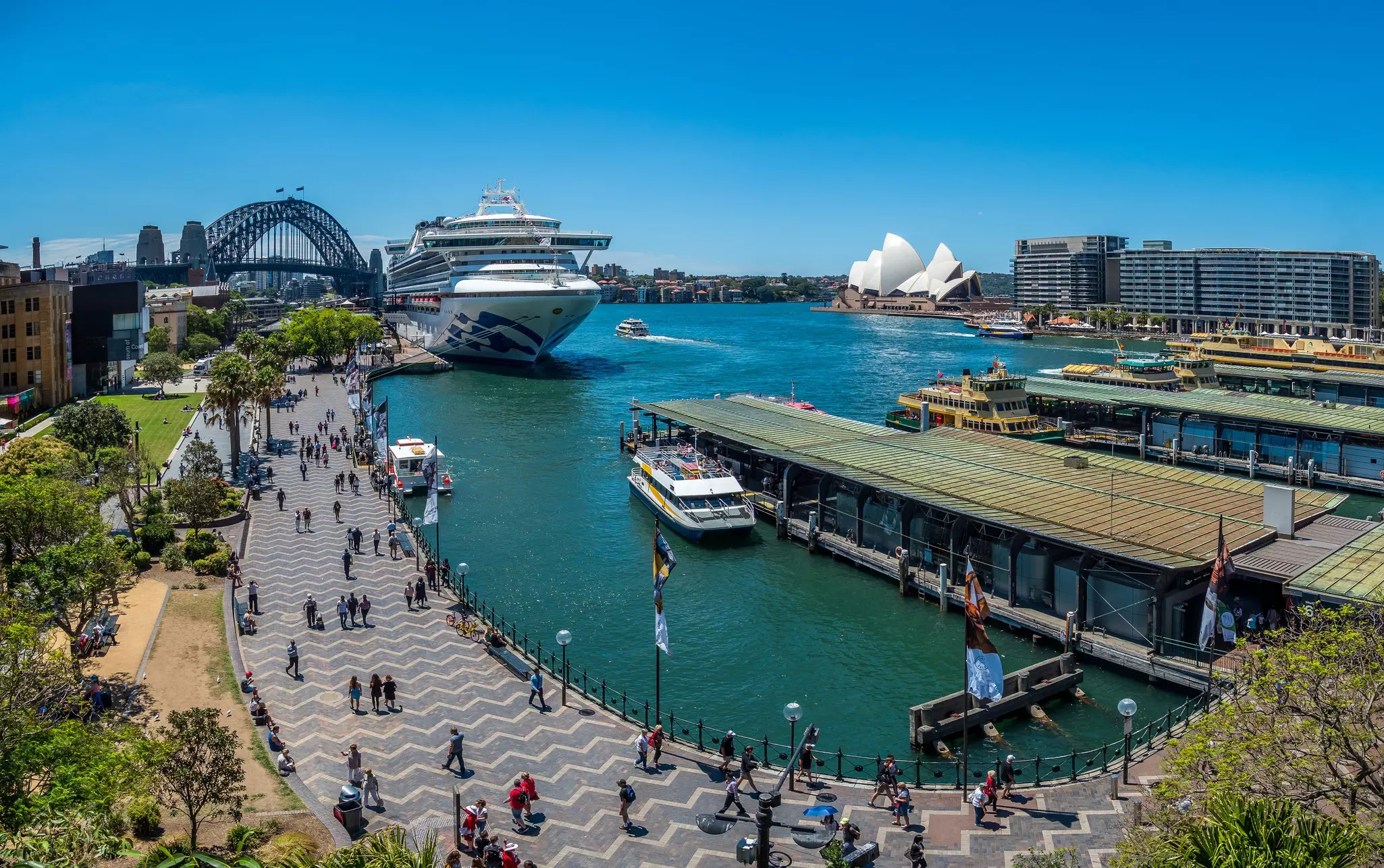 A busy harborfront. A huge cruise ship has docked near a large bridge that looks like a coathanger; a white building with a sail-shaped roof stands on the edge of the quay.