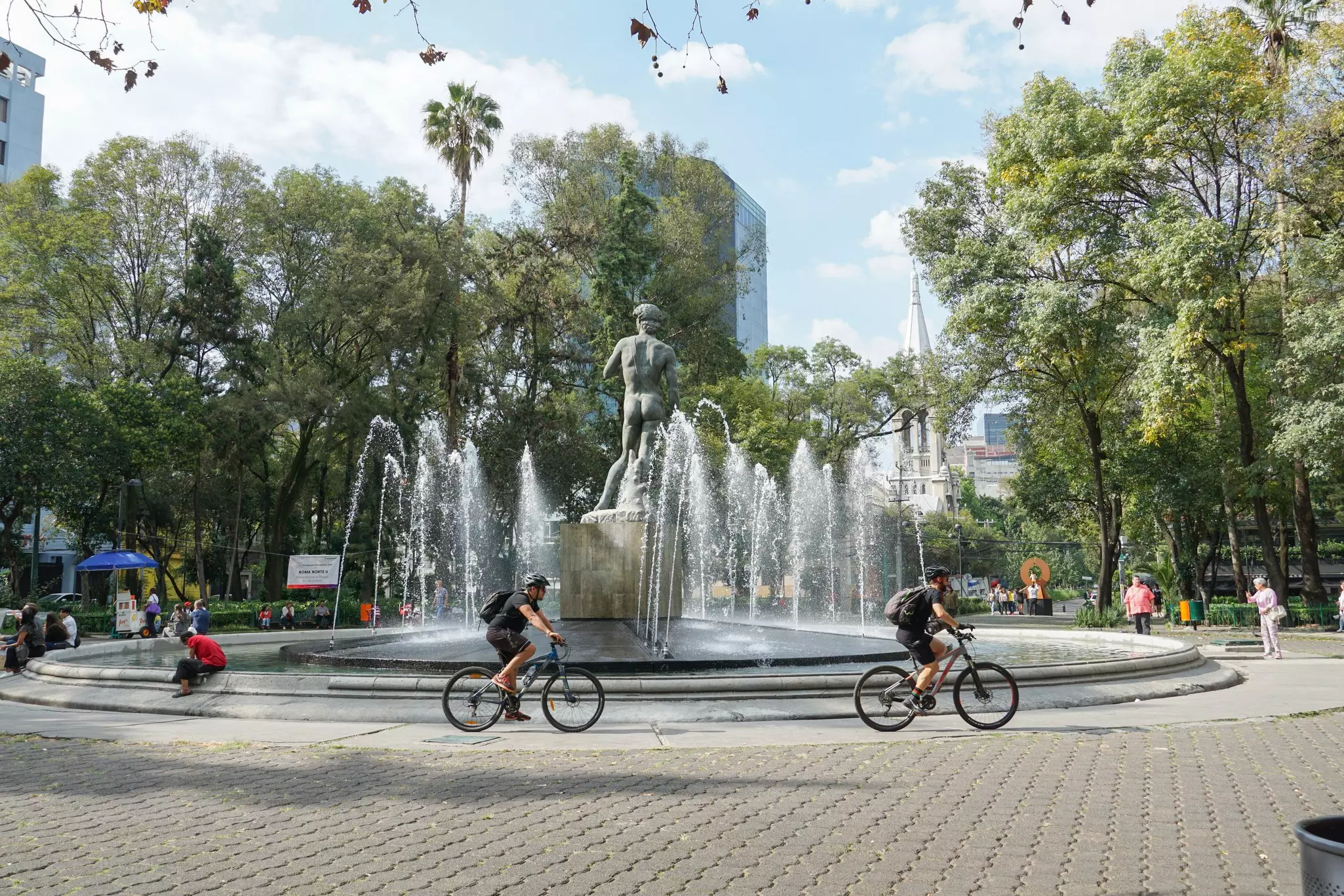 People ride bicycles past a large circular fountain with a replica of Michelangelo's "David" in the center.