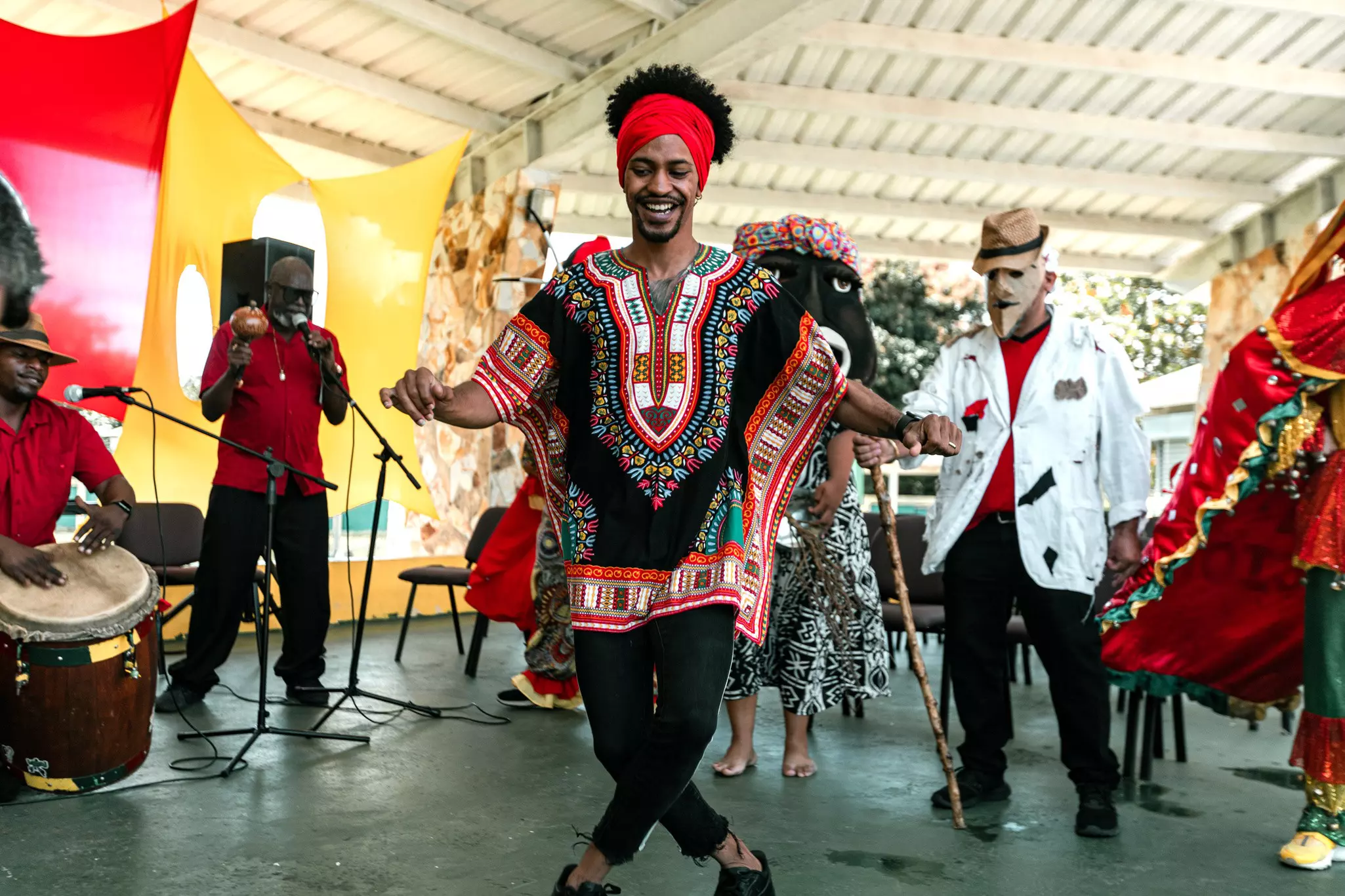 A man wearing a red and black dashiki and a red headwrap dances in front of a band and people wearing traditional masks.