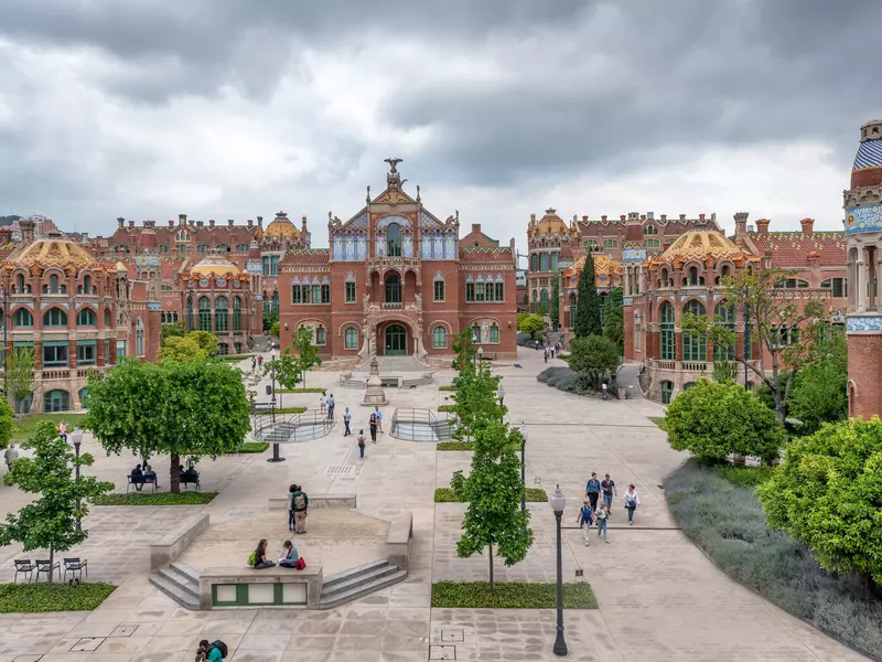 A plaza in Barcelona, Spain, has benches and green trees; it is surrounded by low redbrick buildings with exterior flourishes.