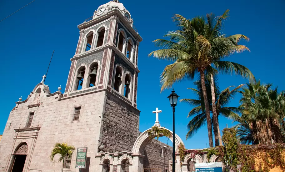 Stone mission church with a tall multi-tiered bell tower and swaying palms; a blue sign reads Mision de Nuestra Senora de Loreto.