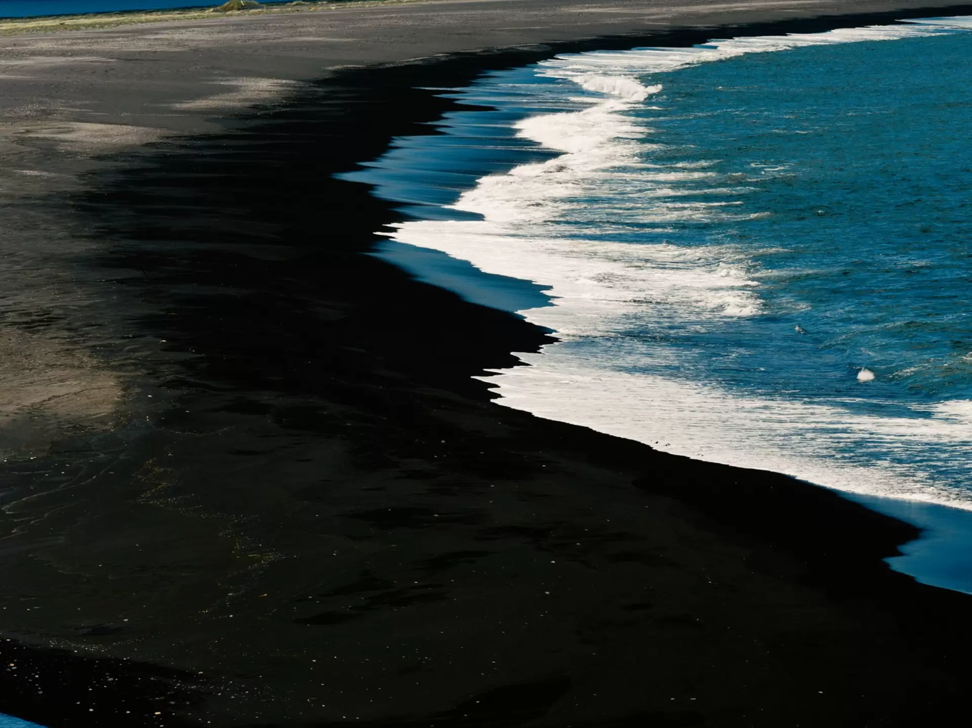 A stark outline of blue waves crashing onto a black-sand beach, viewed from above