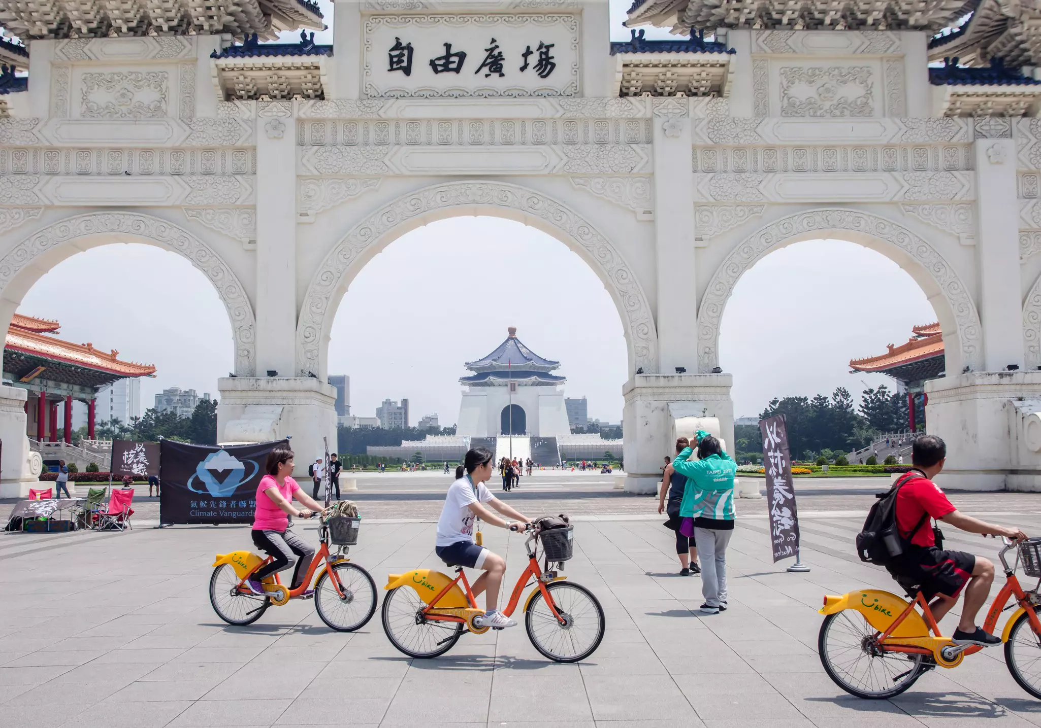 Cyclists on orange-painted bikes pedal past a monumental arch in a city square. A large temple-like mausoleum is seen across the square.