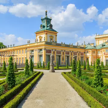 A yellow baroque palace facade behind formal clipped hedges and gravel paths.