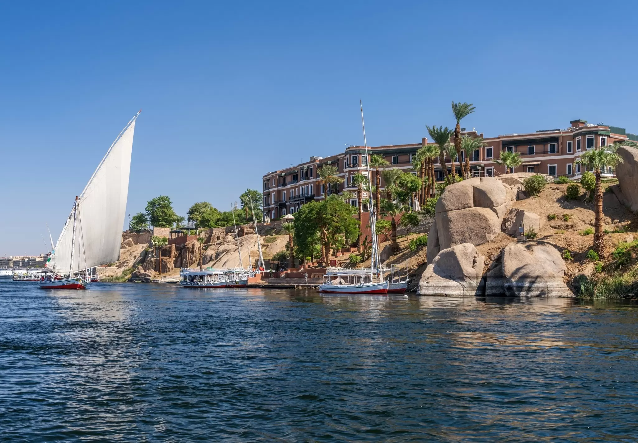 Boats moored near a hotel on a rocky outcrop beside a wide river: the Old Cataract in Aswan, Egypt. 