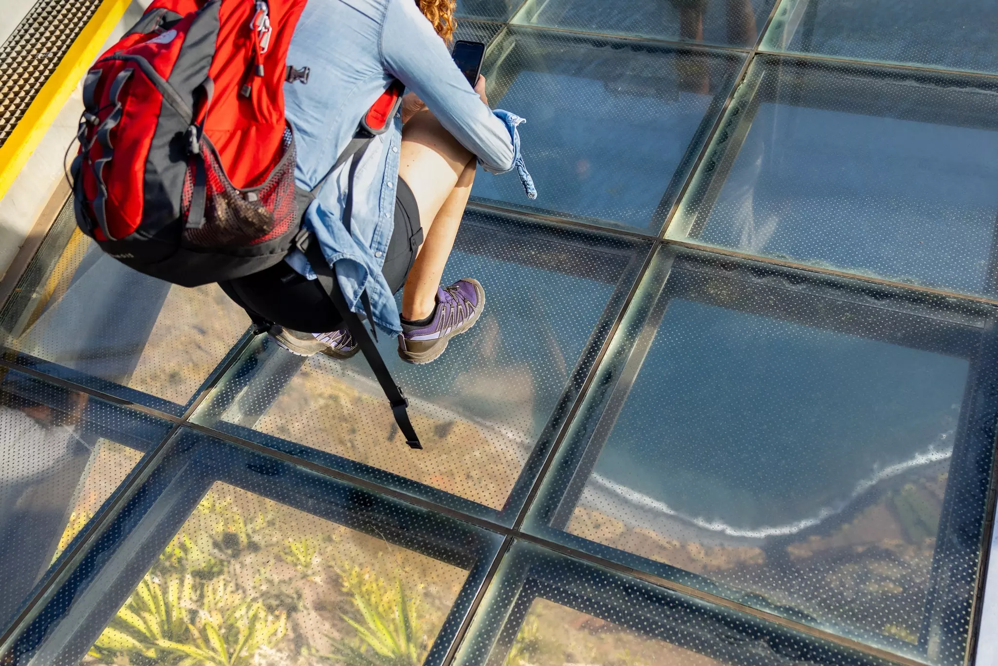 Woman standing on a transparent glass skywalk with water and greenery below.