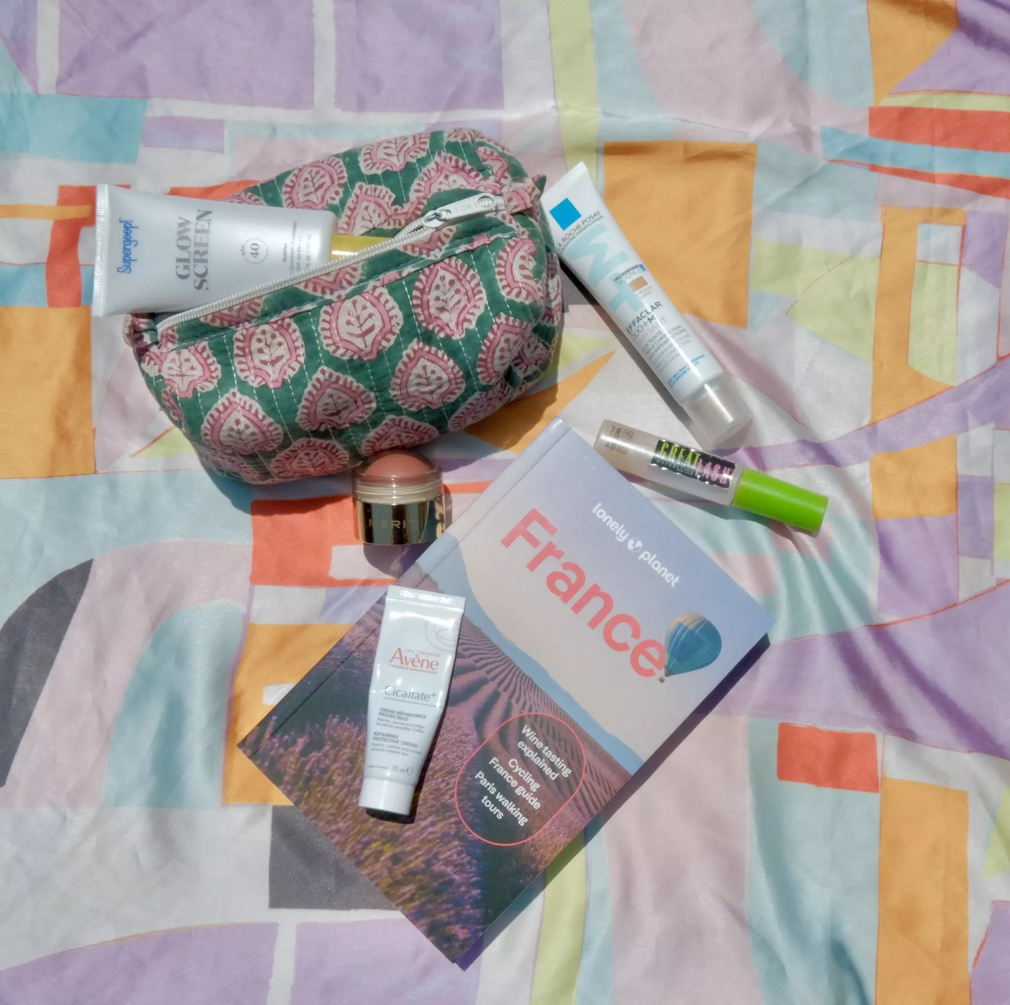A pastel-colored blanket with a guidebook for France and beauty products laid out on it.