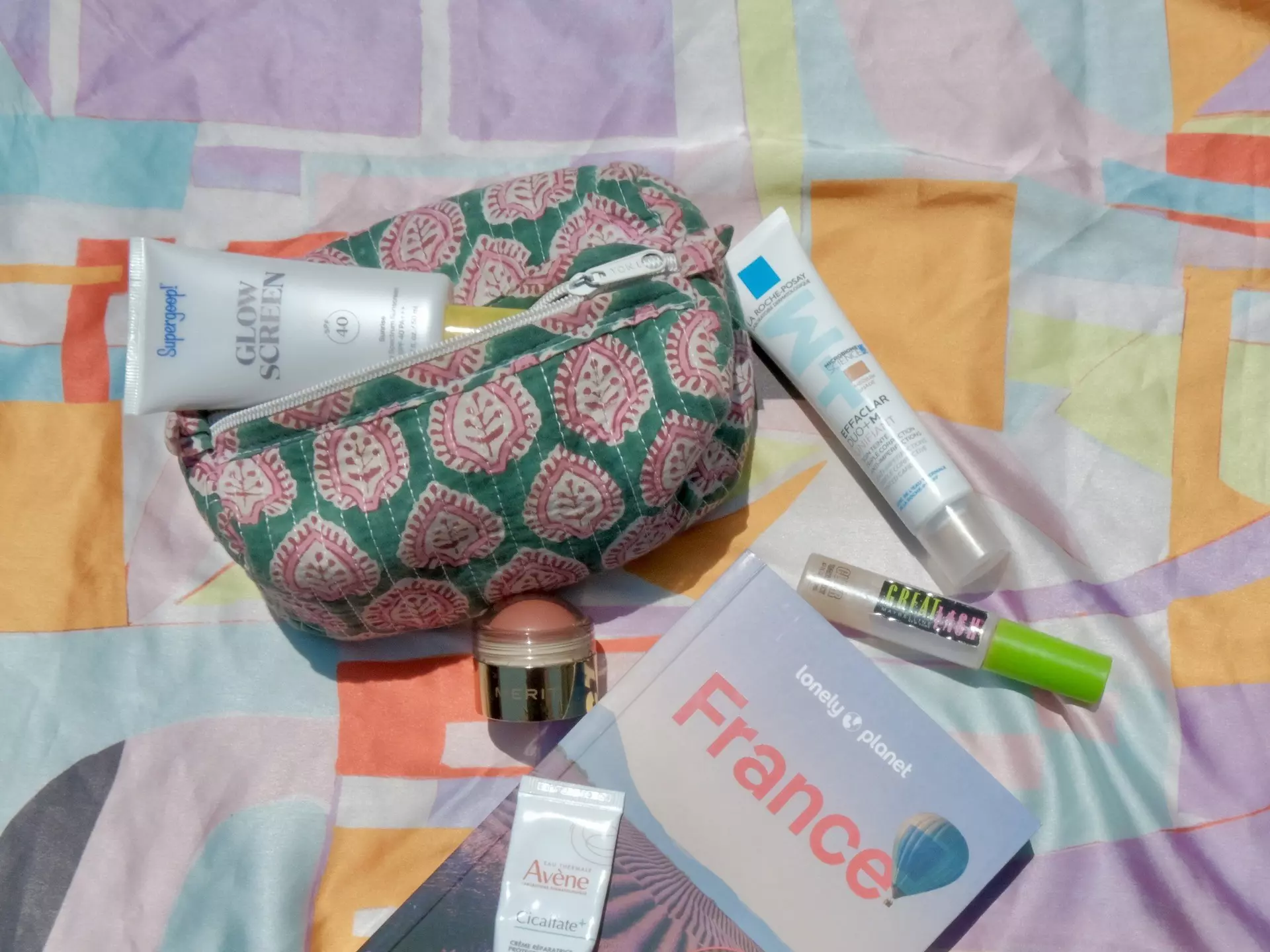A patterned pouch with a zipper on a patterned scarf with a Lonely Planet guidebook for France and makeup products.